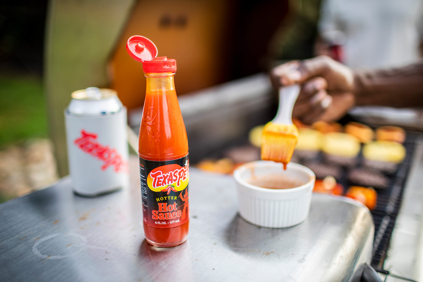 Texas Pete hot sauce bottle on a grill beside canned drink and container of sauce with food brush