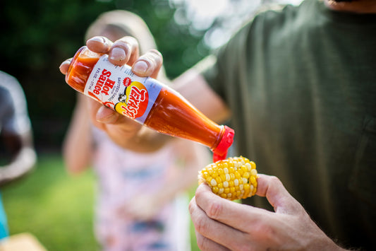 Person pouring Texas Pete sauteed garlic hot sauce onto a corn on the cob with a blurred outdoor background