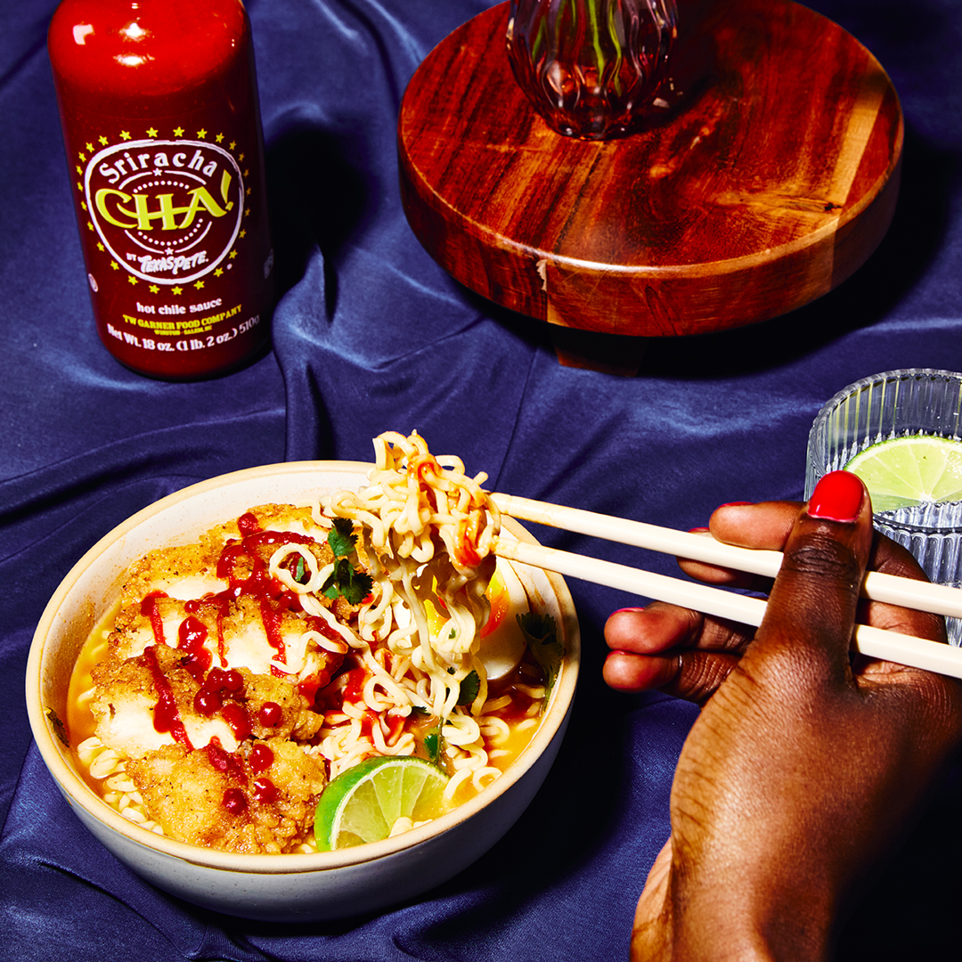 Bowl of ramen with sriracha hot chile CHA sauce by Texas Pete,  and a hand using chopsticks on a blue tablecloth.