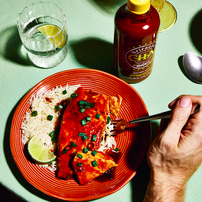 Plate of food with a fork, bottle of sriracha hot chile CHA sauce by Texas Pete, on a green table.
