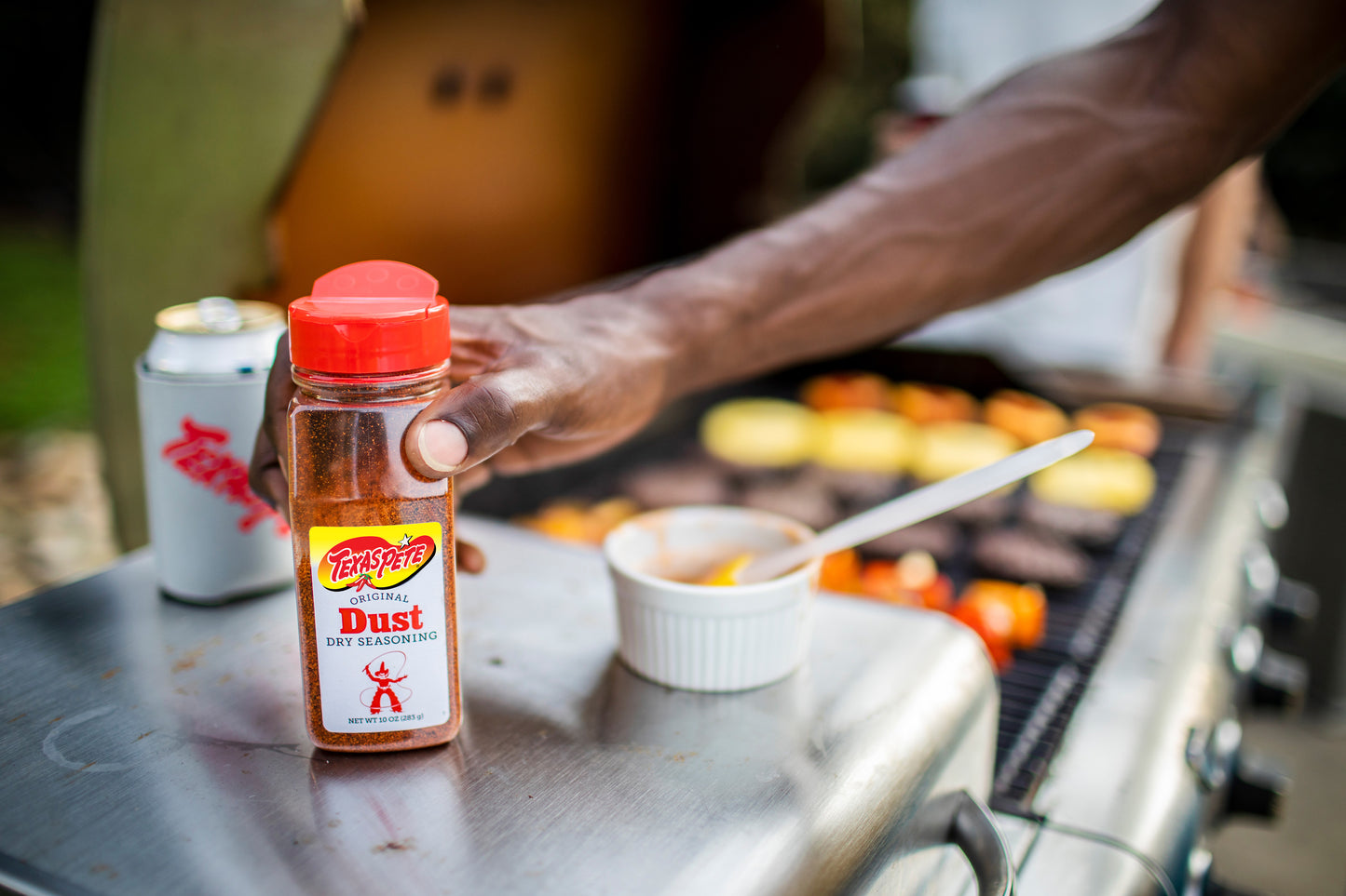 Hand holding a bottle of Texas Pete Original Dust Dry Seasoning over a grill with food.