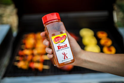 Hand holding a bottle of Texas Pete Original Dust Dry Seasoning with a grill and skewers in the background