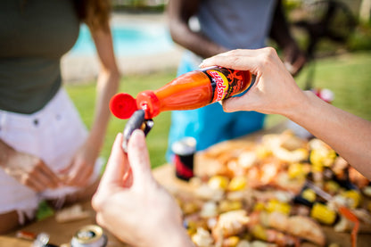 Person holding a bottle of Texas Pete hotter hot sauce over a grill with food in the background