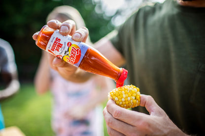 Person pouring Texas Pete sauteed garlic hot sauce onto a corn on the cob with a blurred outdoor background