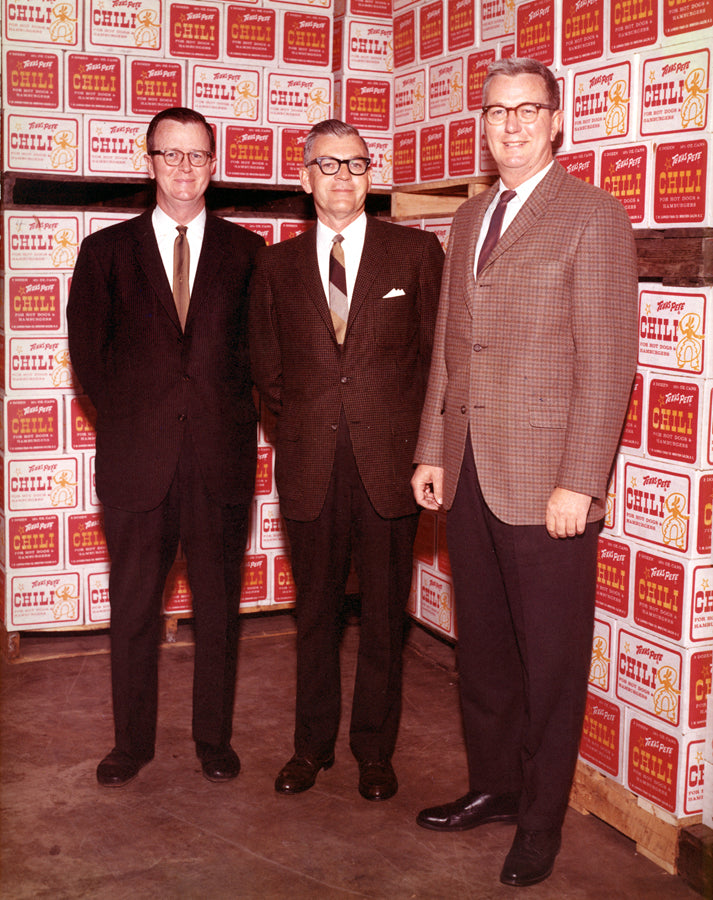 Brothers Ralph C. Garner, Harold H. Garner Sr., and Thad W. Garner standing in front of a wall of chili canisters.